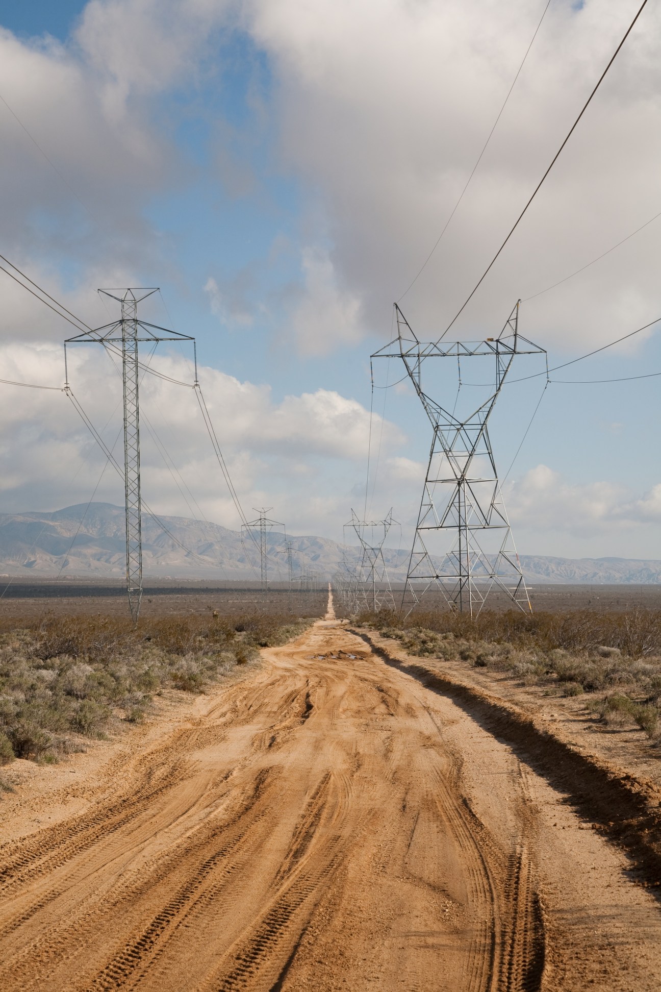 High-voltage power transmission towers and lines stretching across arid desert landscape with mountains in the distance.