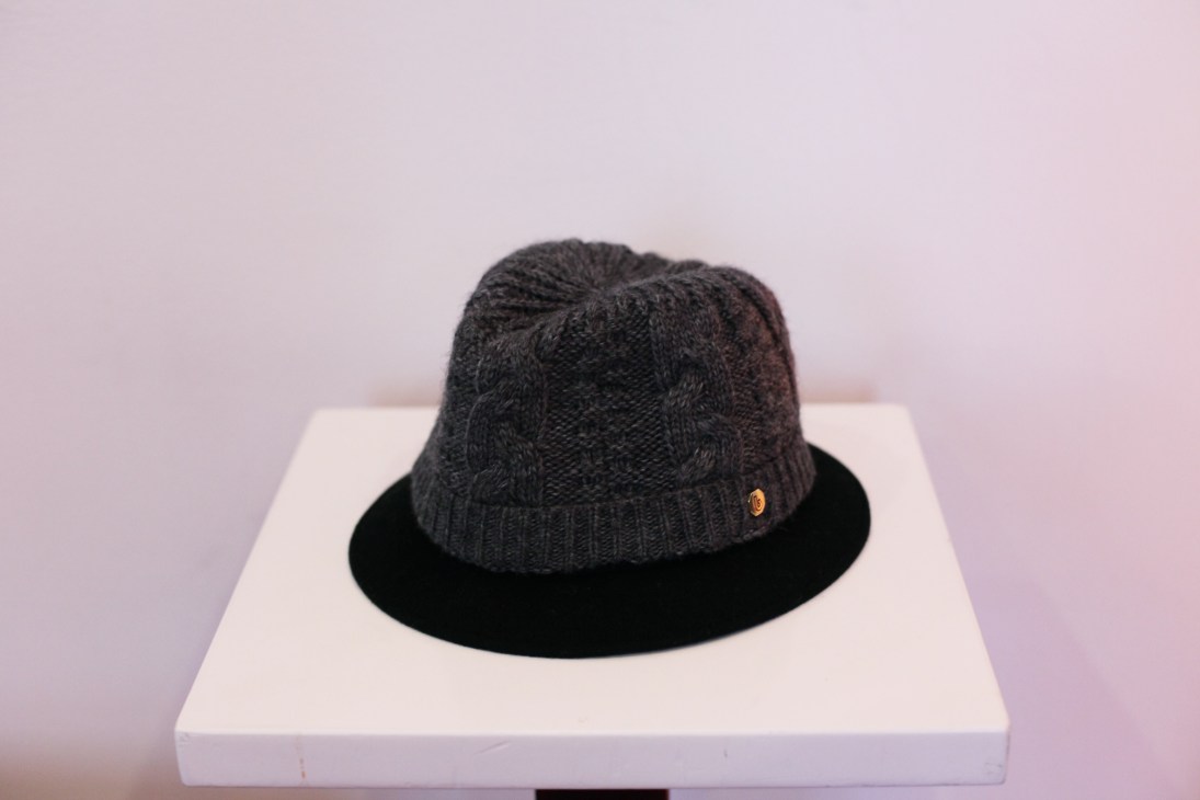 Black knitted wool hat with cable knit pattern and small gold badge displayed on white block.