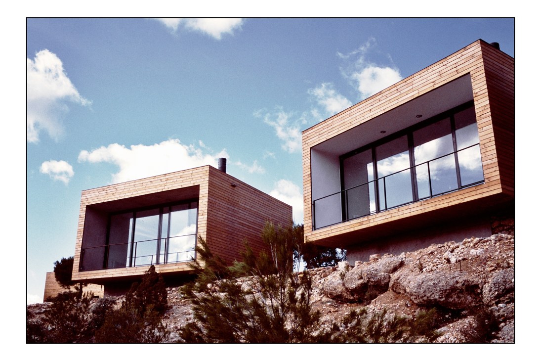 Two modern wooden box-shaped rooms with large windows perched on a rocky hillside under blue sky.