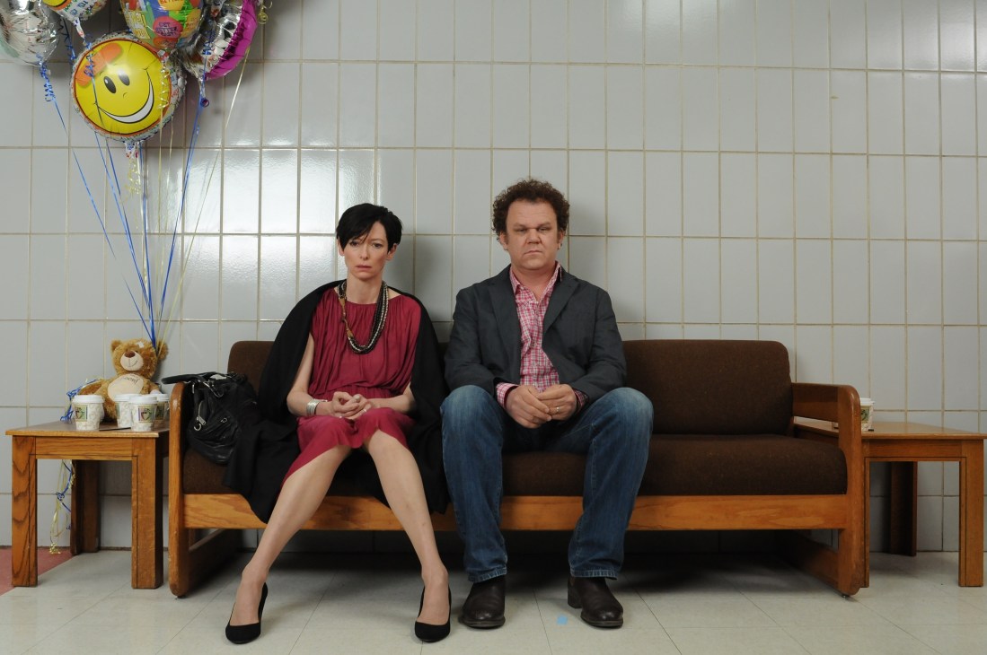 Two people sitting on a brown couch against a white tiled wall with colorful balloons beside them.