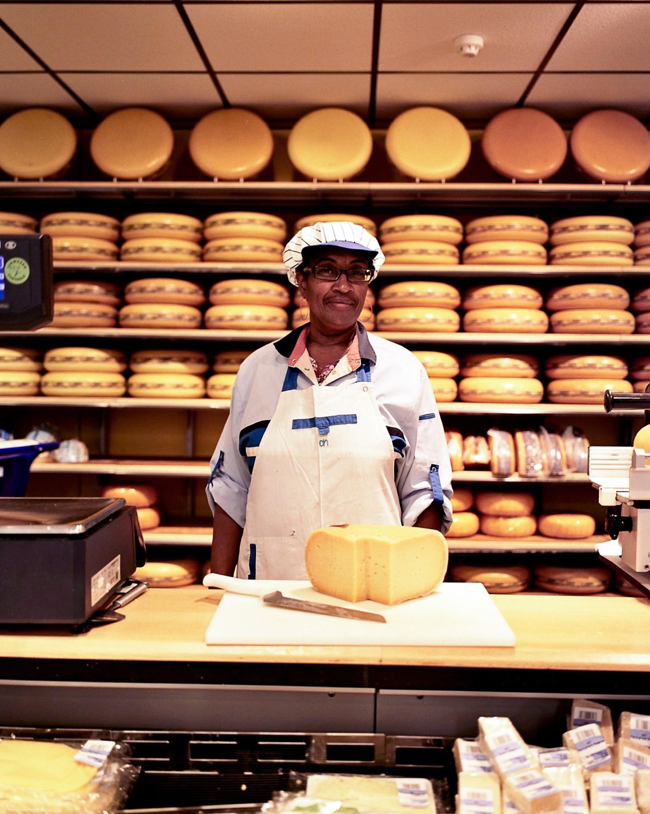 Cheesemonger at Albert Heijn store counter with wheels of cheese and shelves of cheese wheels in background.