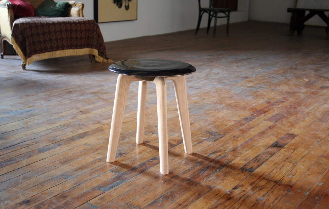 Modern stool with black ceramic seat and light ash wood legs on wooden floor.