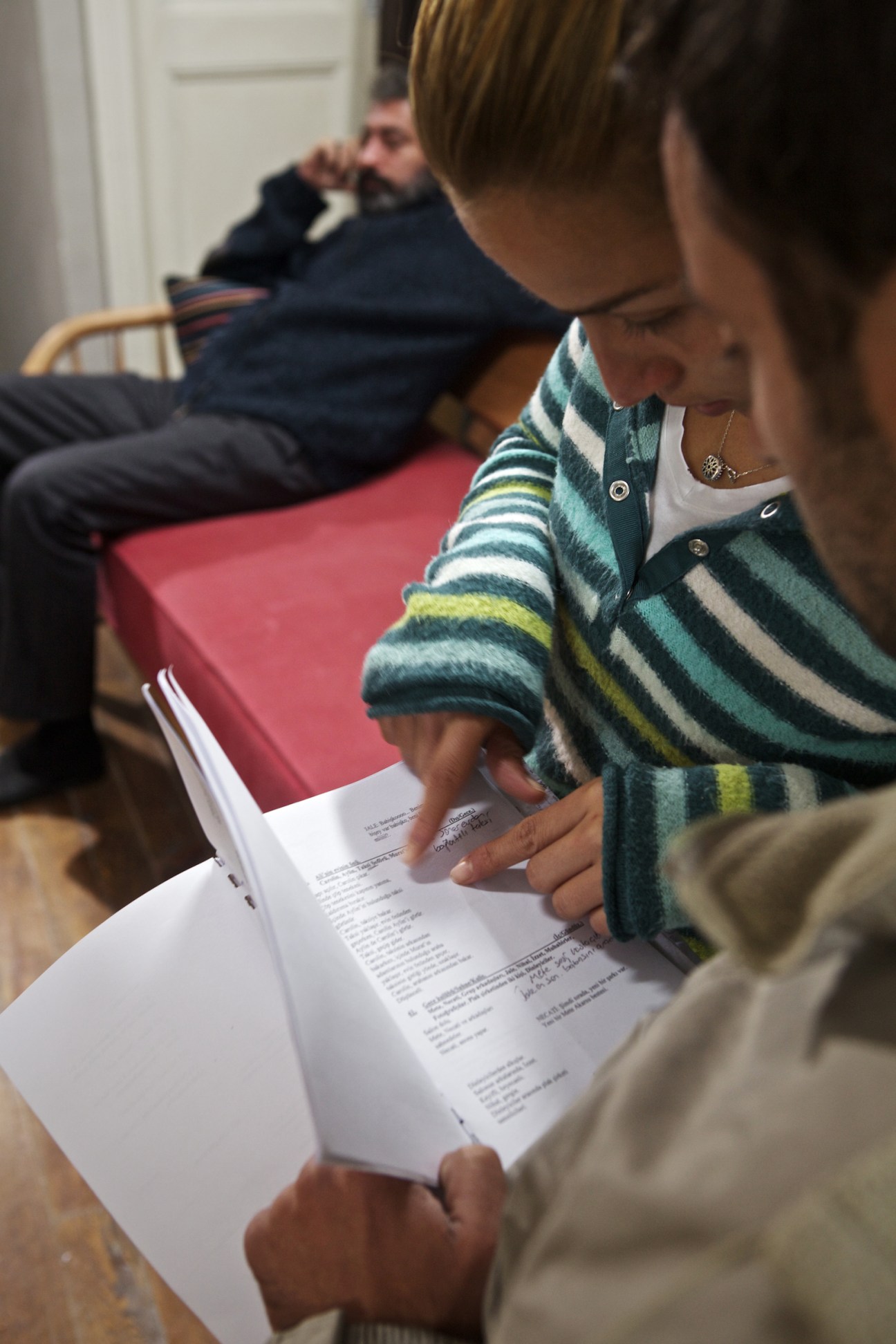 Woman in striped cardigan reads script pages on a film set while man sits in background.