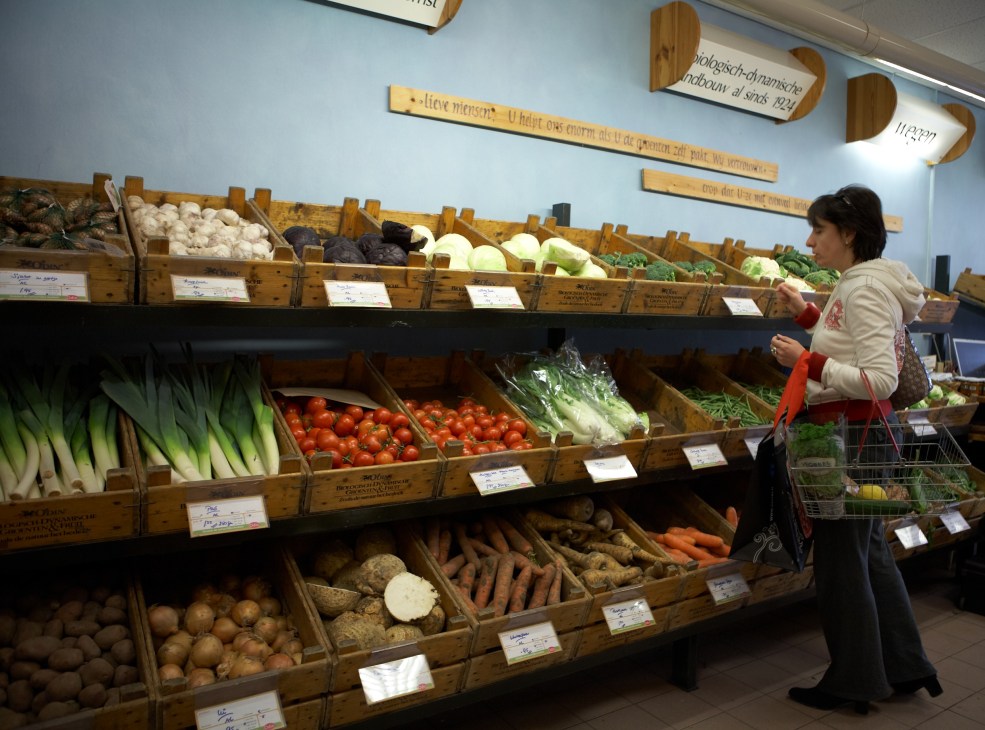 Customer shopping at wooden produce displays in Gaia supermarket with fresh vegetables and fruits.