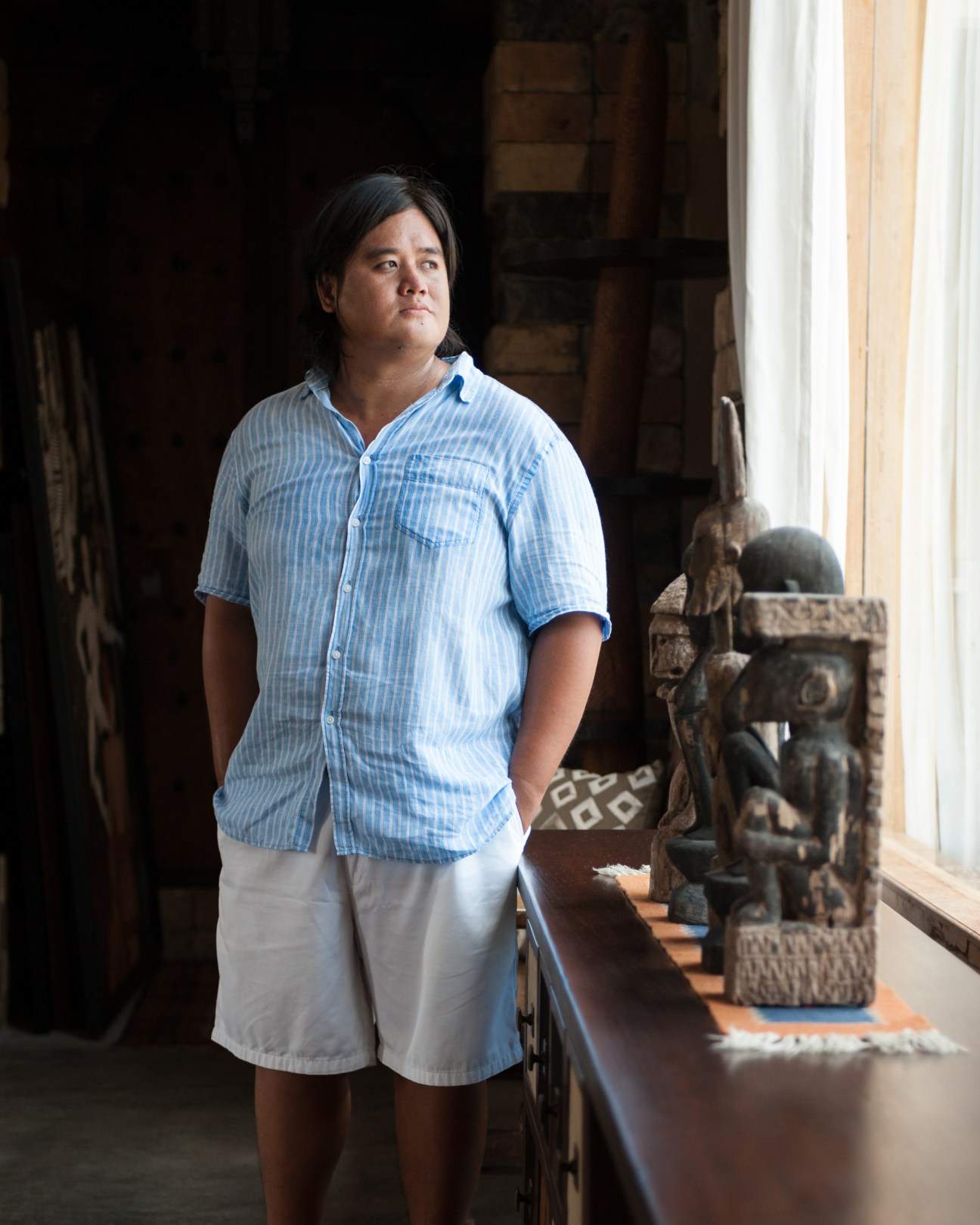 Karl Aguila stands in his workshop wearing a blue striped shirt, surrounded by wooden furniture and sculptures.