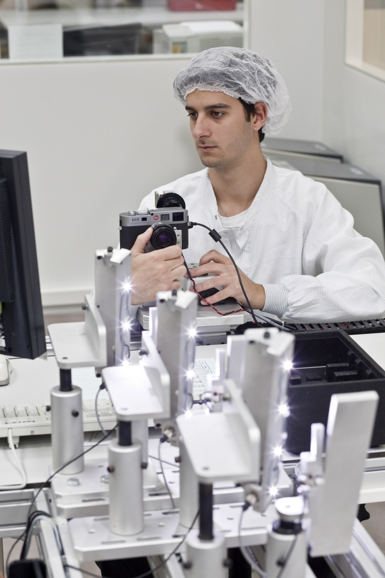 Man in white lab coat and hairnet holds Leica camera at workstation with LED-lit equipment.