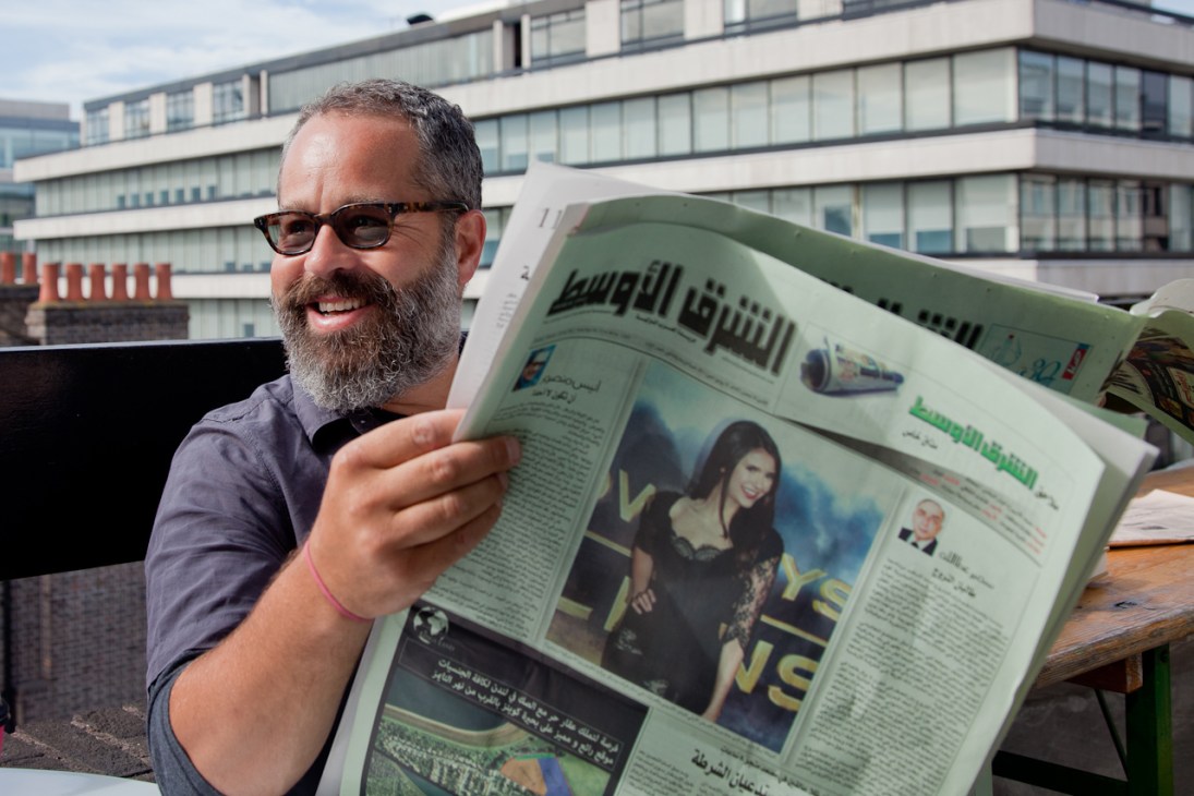Man with beard and glasses holding open an Arabic-language newspaper on a rooftop, smiling at the camera.