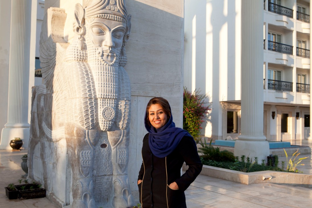 Woman in hijab stands before a large white stone statue of a bearded Persian figure with wings at the Dariush hotel.