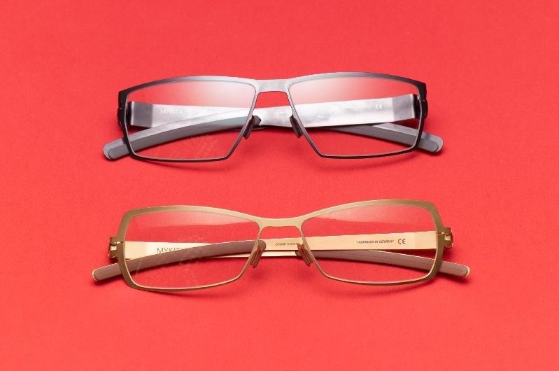 Two eyeglass frames displayed on a red background: one black rectangular frame and one gold rectangular frame.