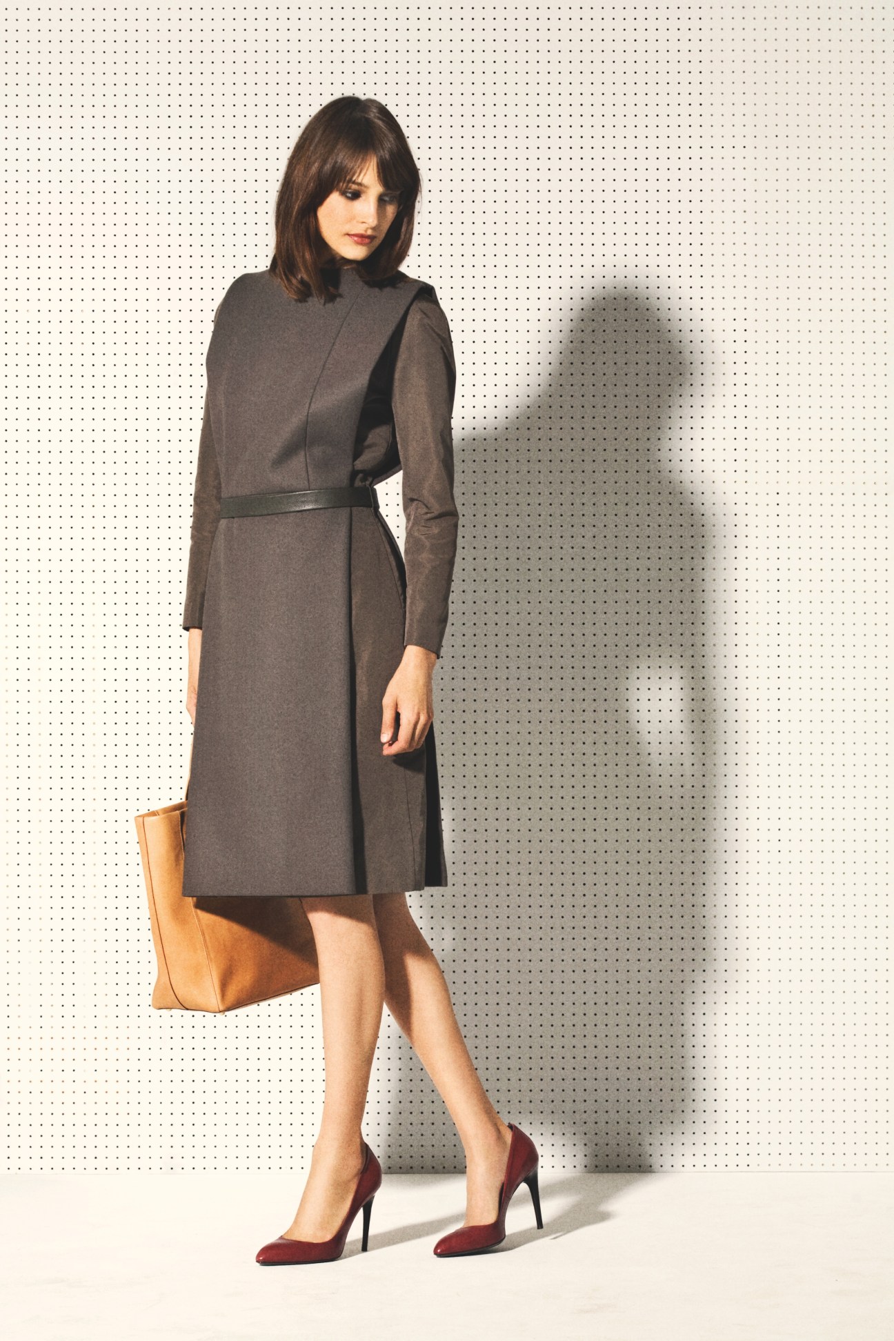 Woman wearing a charcoal faille and wool dress with strong pleats by Joaquin Trias, paired with burgundy heels and a tan leather bag.