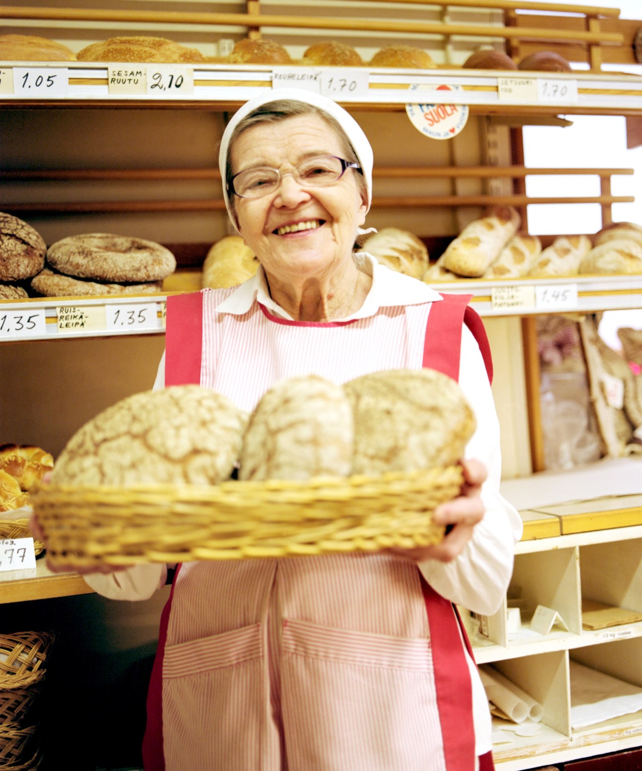 Baker Elvi Avikainen holds a basket of bread in her bakery with shelves of loaves behind her.