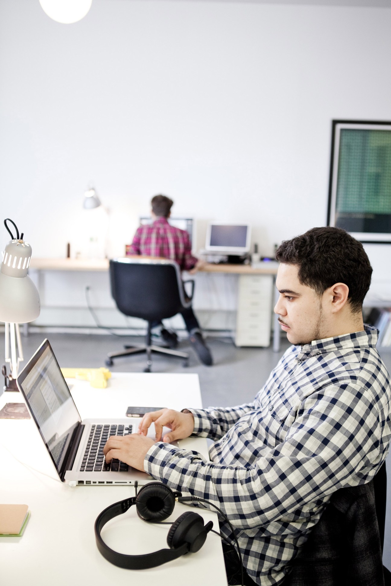 Man in checkered shirt working on laptop at desk with headphones nearby, colleague in background.