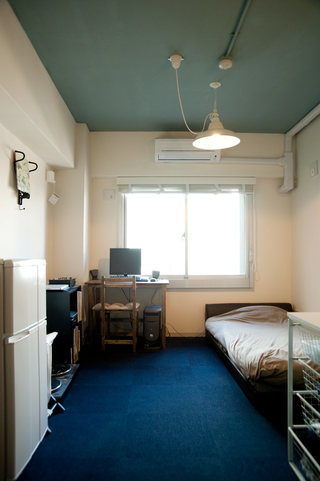 A compact studio room with a bed, desk, window, and minimal furnishings featuring a teal ceiling.