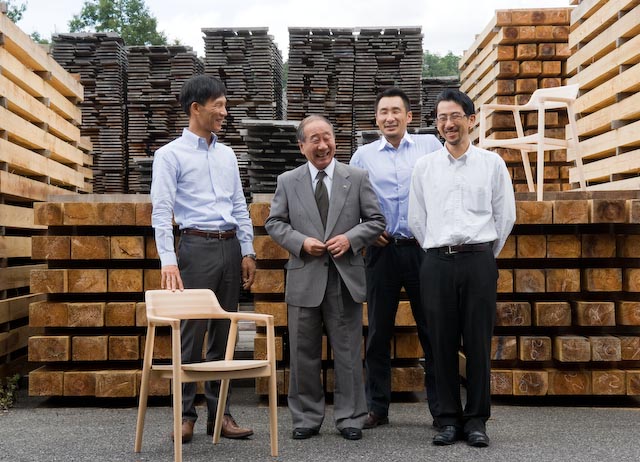Four men in business attire stand among stacked wood at a furniture factory, with a wooden chair in the foreground.