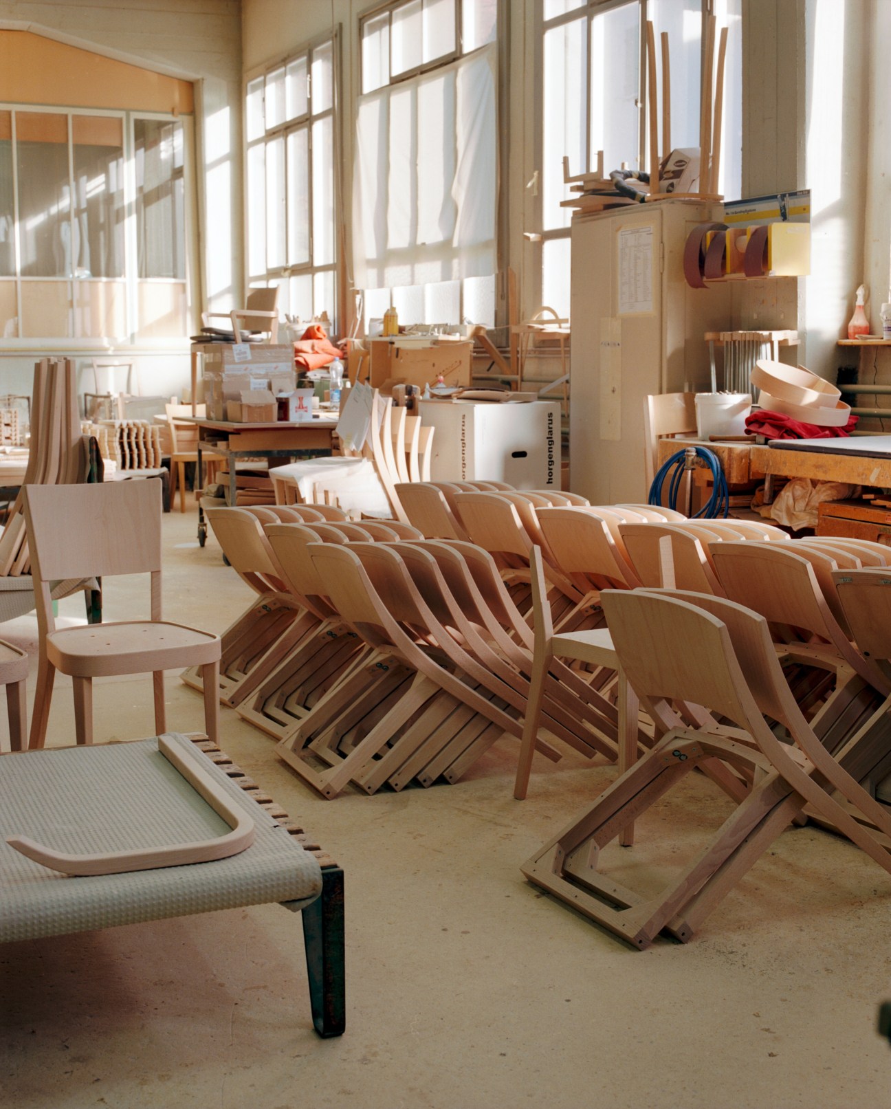Unfinished wooden Lyra chairs stacked in a bright factory workshop with tools and materials.