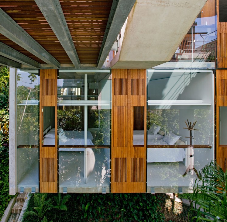 Guest room with large windows framed by wooden slats, showing bedrooms inside and surrounding forest vegetation.