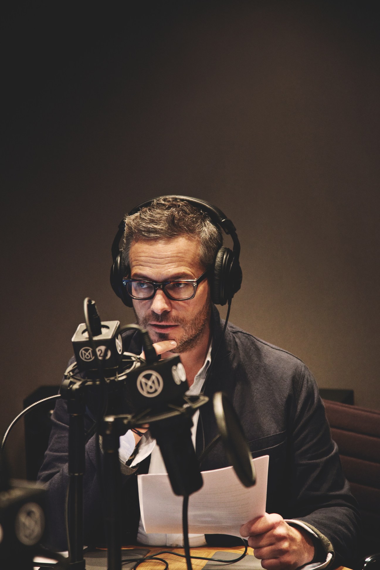 Tyler Brûlé wearing headphones and glasses speaks into a studio microphone while holding papers.