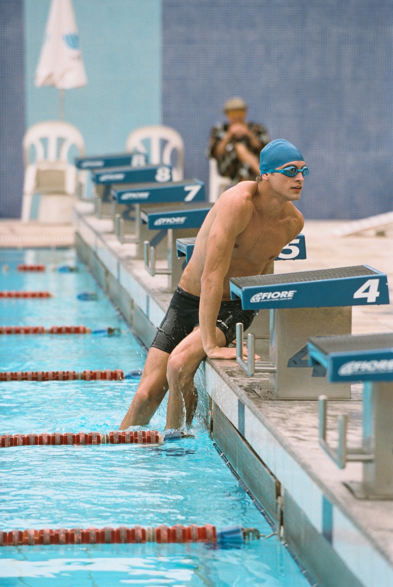 Swimmer in blue cap and Asics goggles poised on diving board at pool