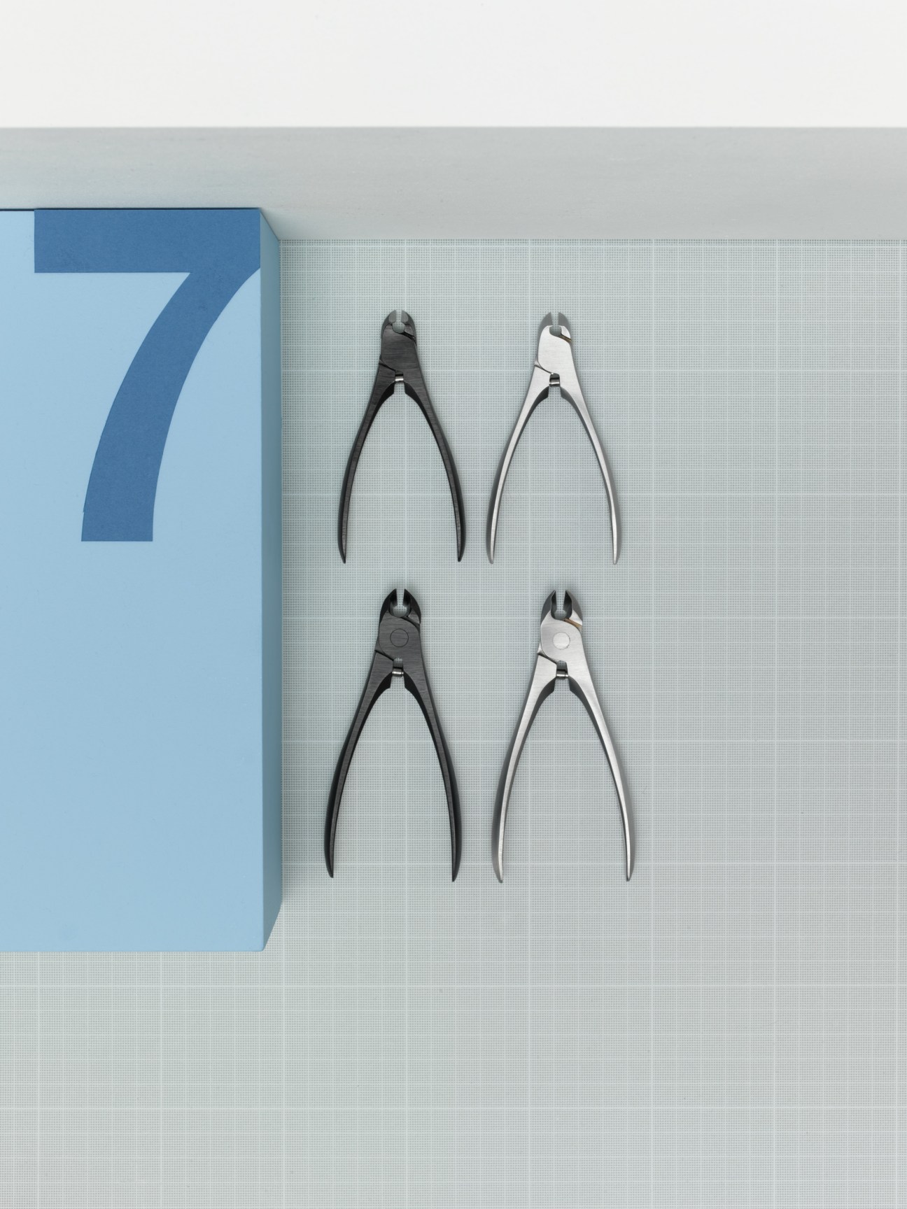 Four Suwada handmade nail clippers in black and silver arranged on a light grid background next to a blue box.