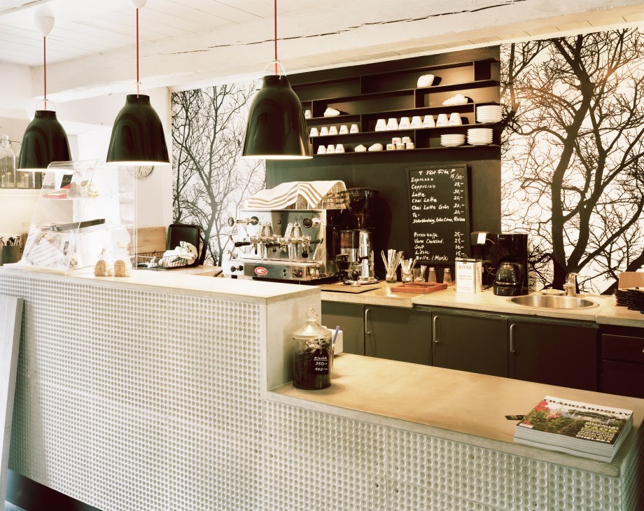 Modern café counter with green pendant lights, espresso machine, white cups on shelves, and bare tree branches visible through windows.