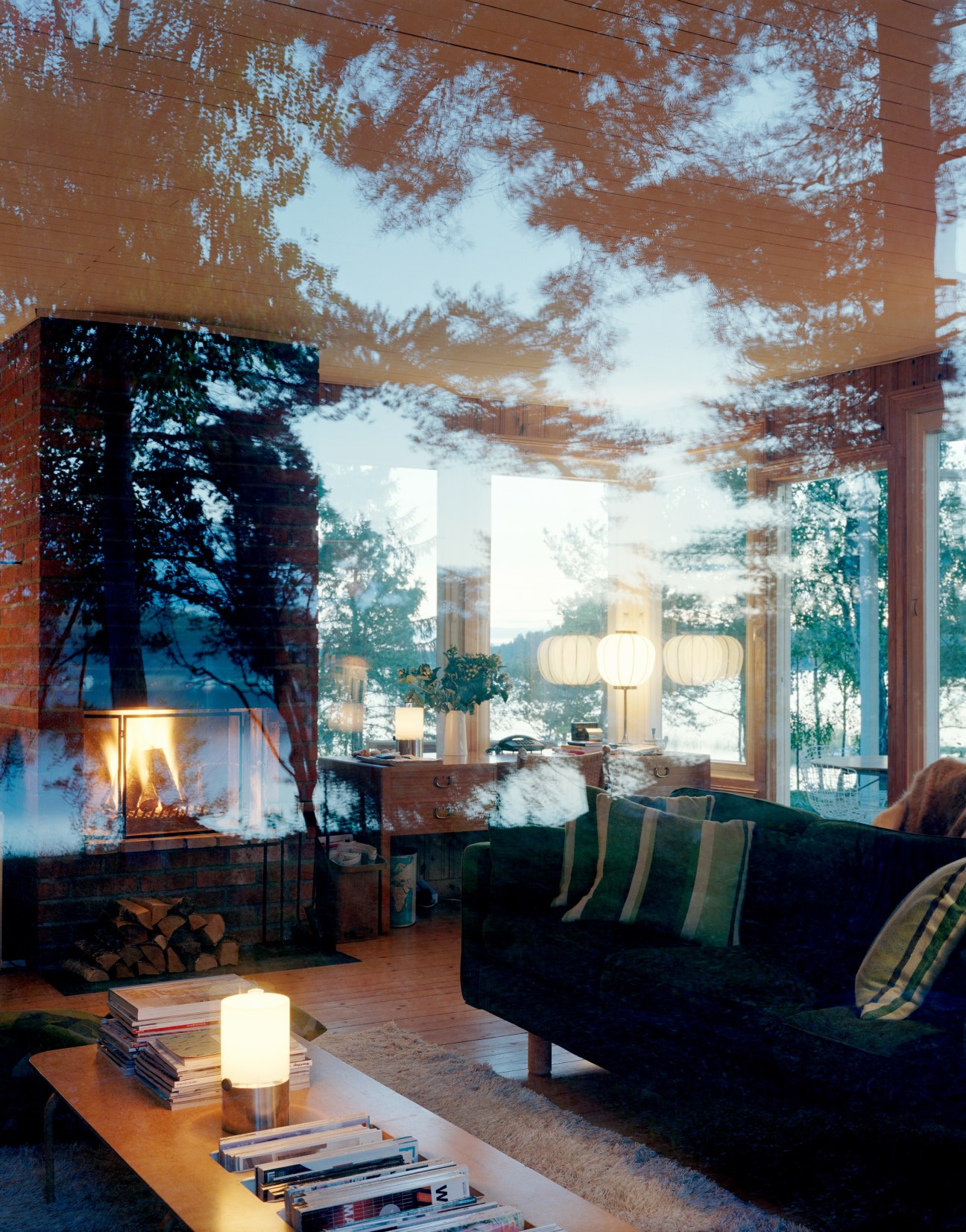 Living room interior with fireplace, moss green sofa, desk, and large windows overlooking trees at dusk.
