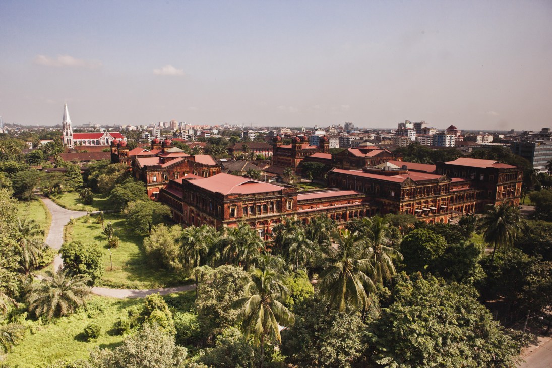 Aerial view of the red-brick Secretariat complex in Rangoon surrounded by lush green gardens and palm trees.