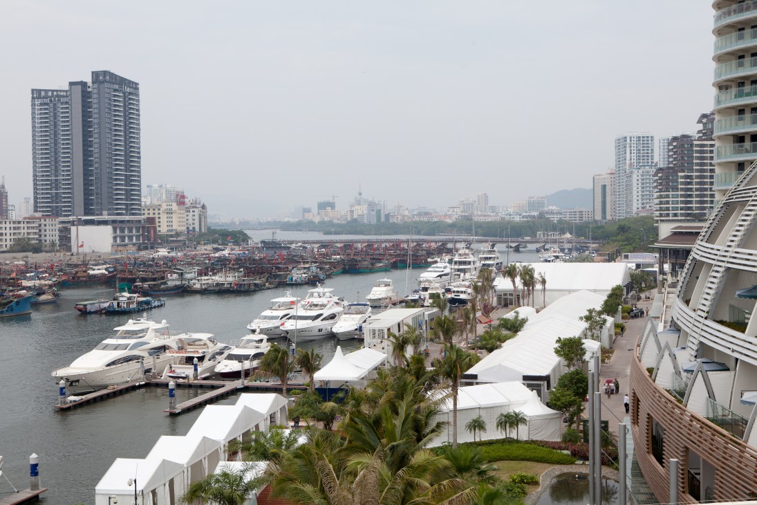 Luxury yachts docked at a marina with modern high-rise residential buildings and palm trees in Sanya, China.