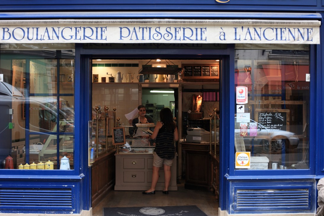 Blue-fronted bakery shopfront with 'Boulangerie Patisserie à l'Ancienne' signage and customer at counter