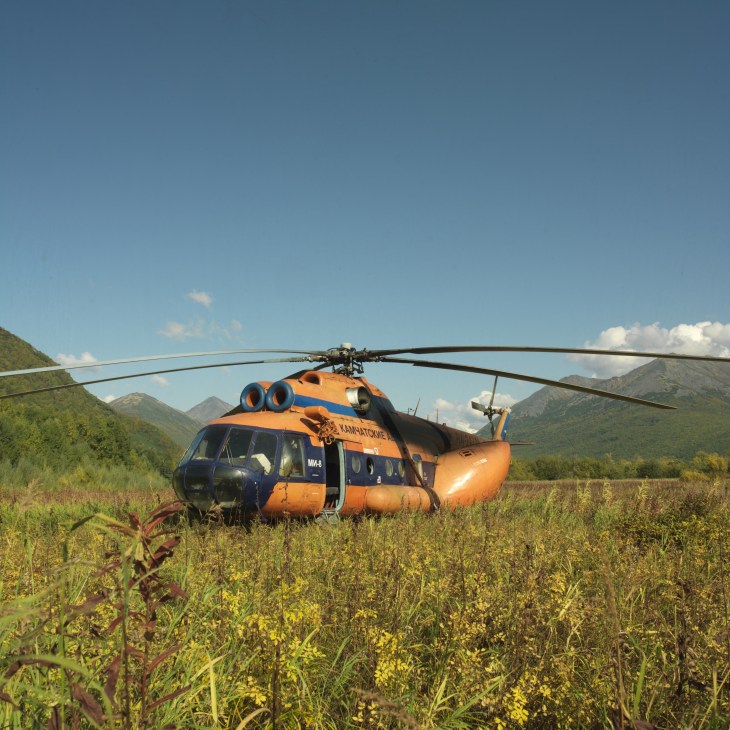 An orange and blue Mi-8 helicopter lands in a grassy valley surrounded by mountains in Kamchatka.
