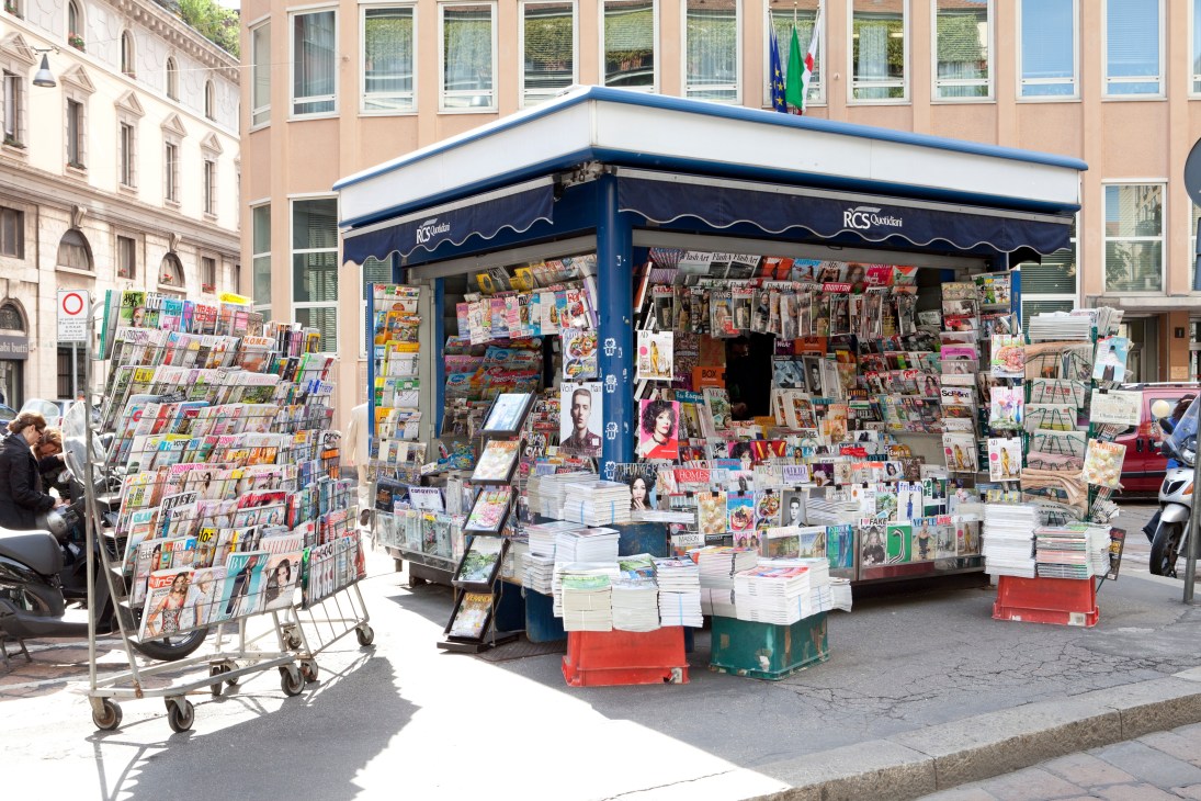 A blue newsstand kiosk on a Milan street corner displaying magazines, newspapers and publications on multiple racks and surfaces.