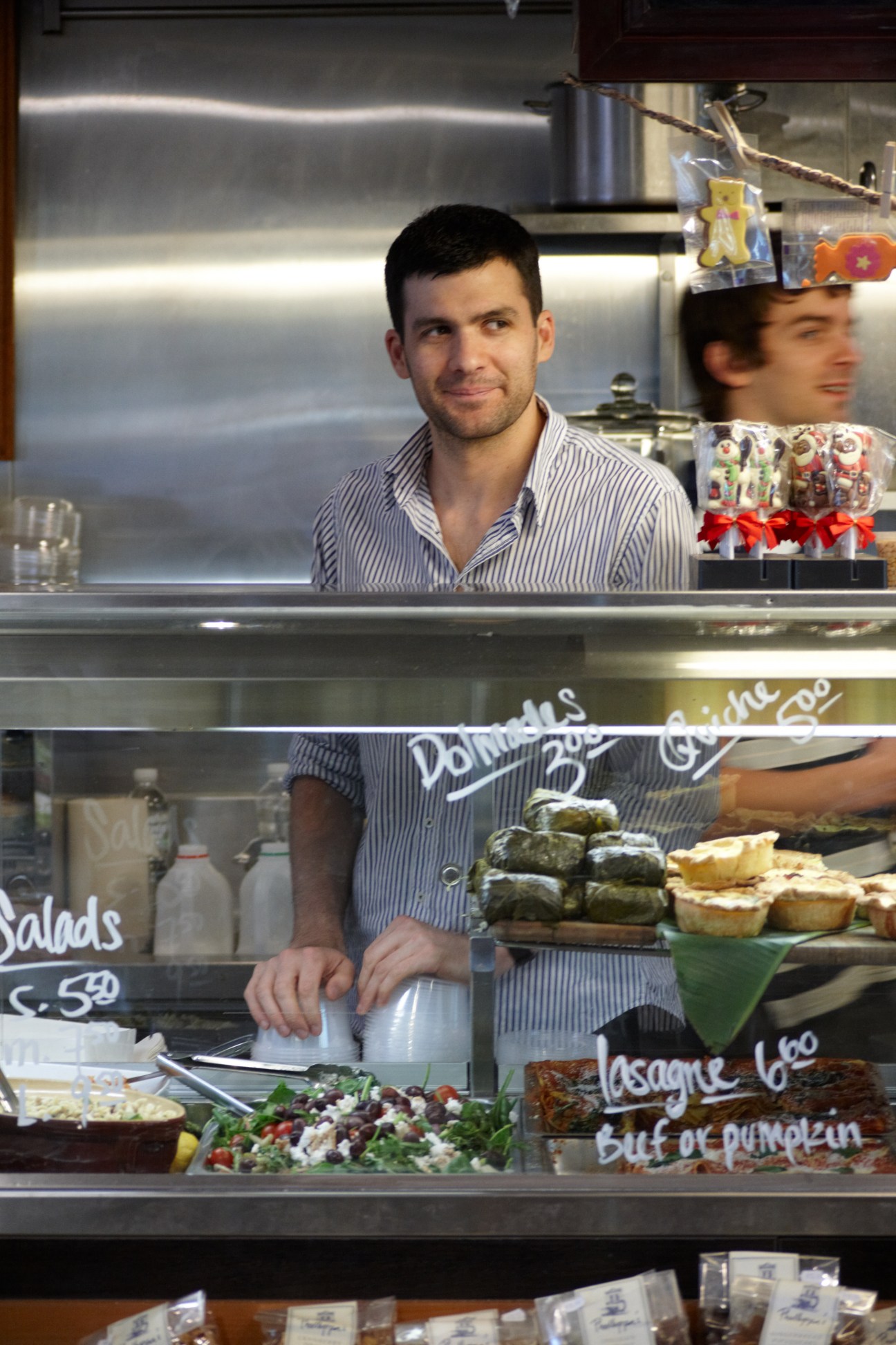 A man in a striped shirt stands behind the counter at Bottega del Vino, a deli with display cases of food items.
