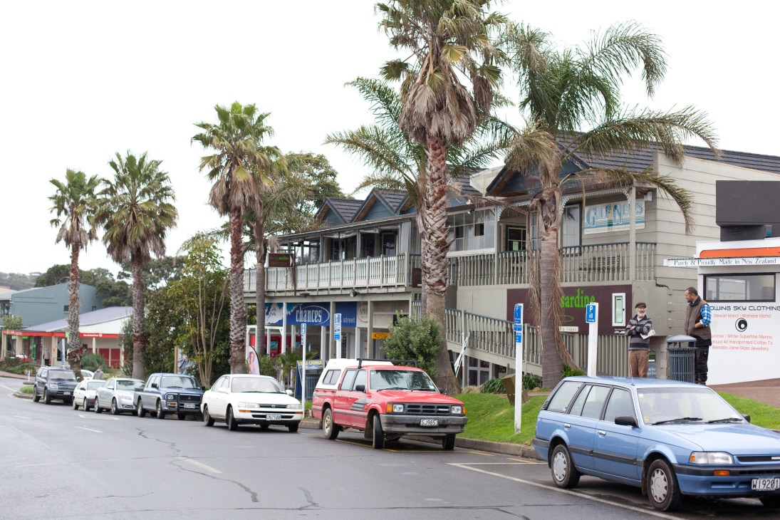Street view of Oneroa village showing colonial-style buildings, palm trees, parked cars and storefronts.