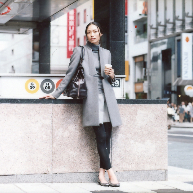 Woman in grey Stella McCartney coat and Drawer jumper standing against concrete wall in urban setting.