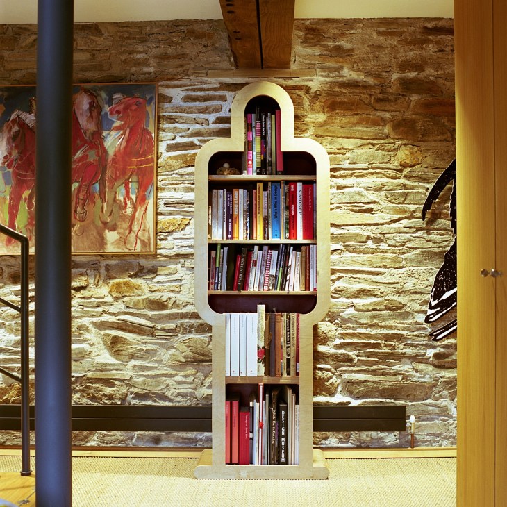 A man-shaped wooden bookcase with cream-colored frame and dark shelves filled with colorful books against a stone wall.