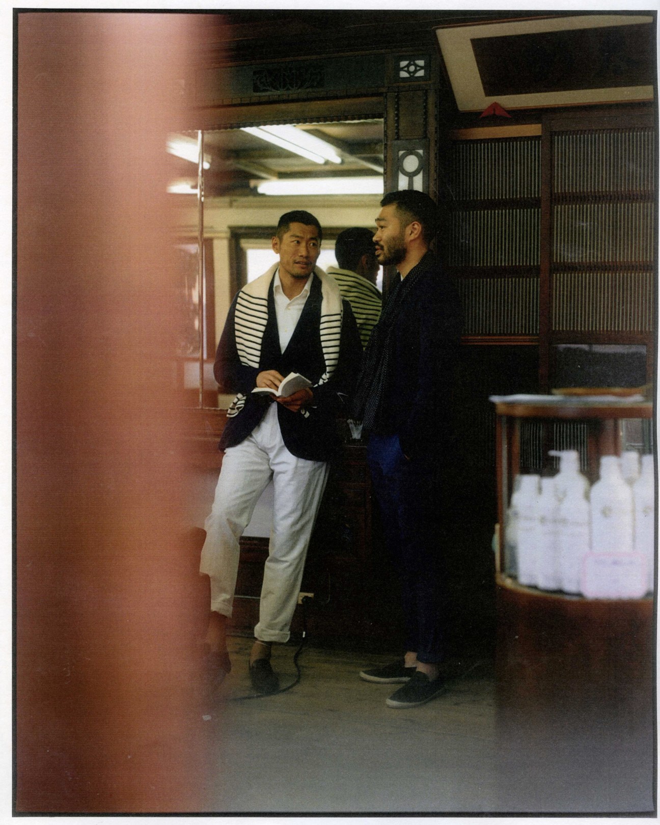 Two men stand in a traditional Japanese barber shop, one wearing a striped cardigan and white trousers, the other in dark clothing.