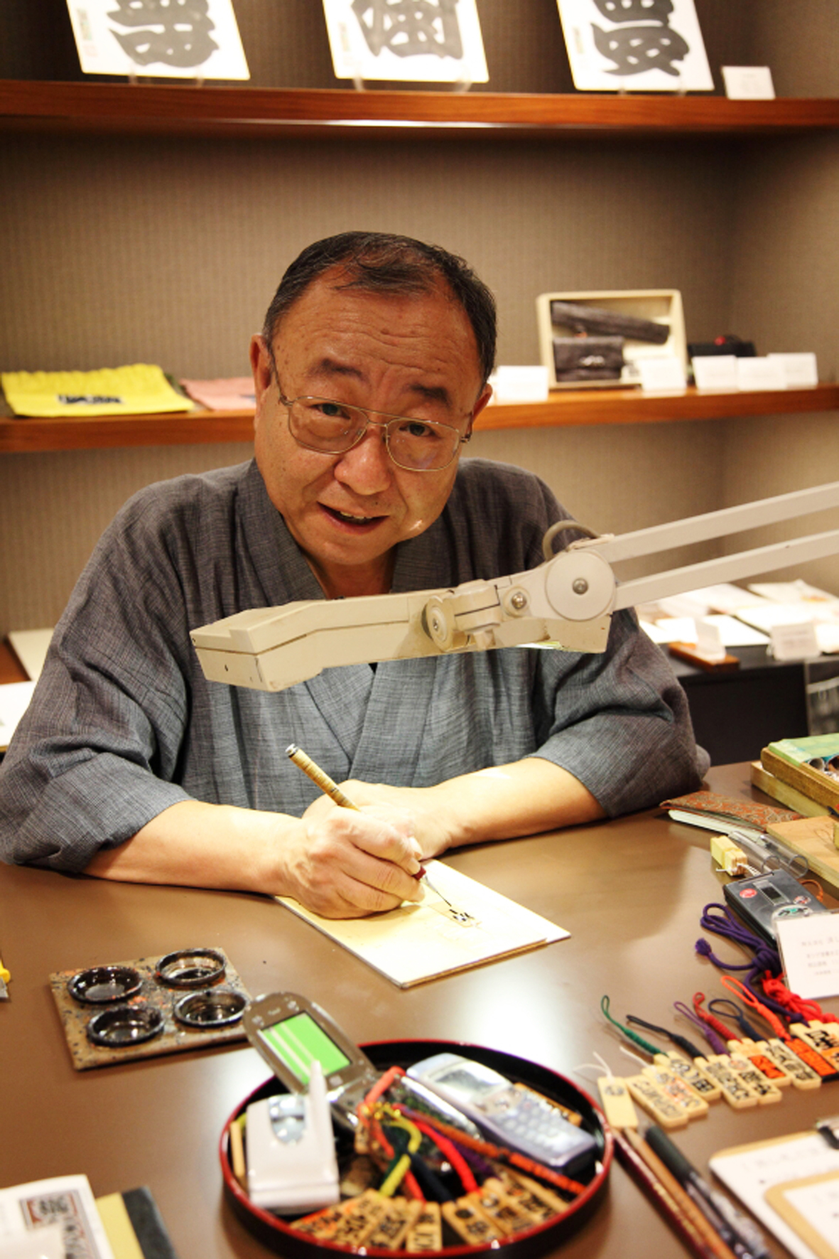 An older man with glasses works at a desk with a desk lamp, drawing or writing on paper surrounded by electronic components and tools.