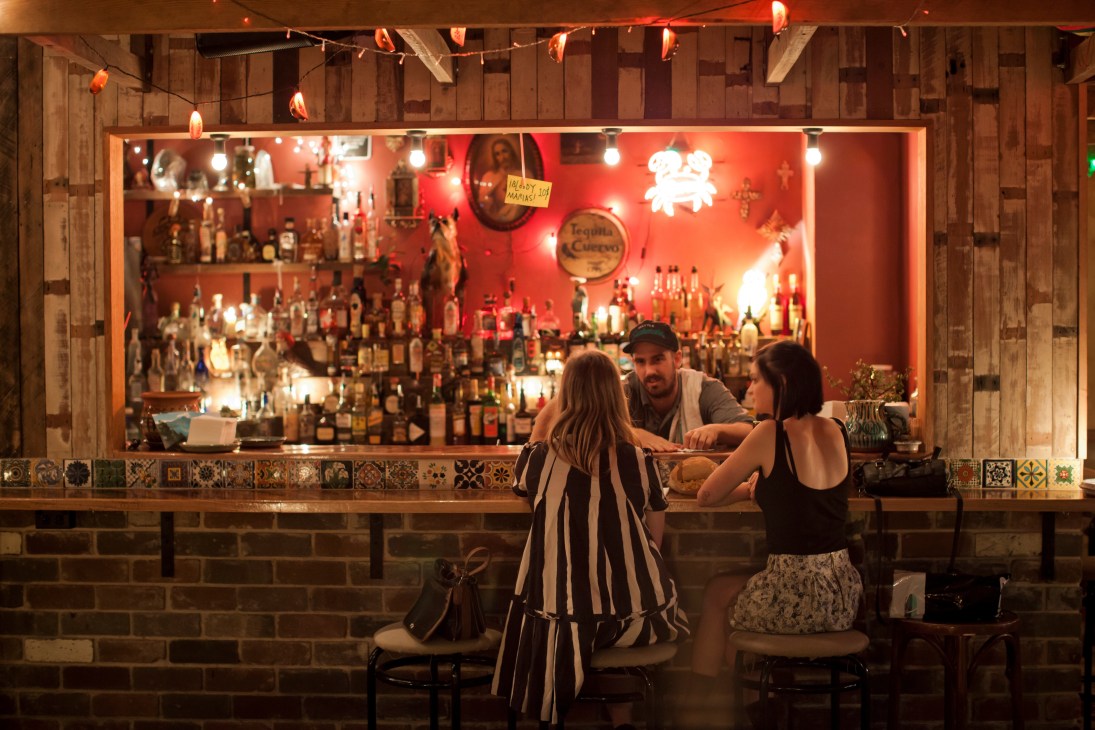 Two customers sit at the bar of Tio's Cerveceria, a tequila bar in Sydney with a red-lit backdrop and numerous bottles displayed behind the counter.