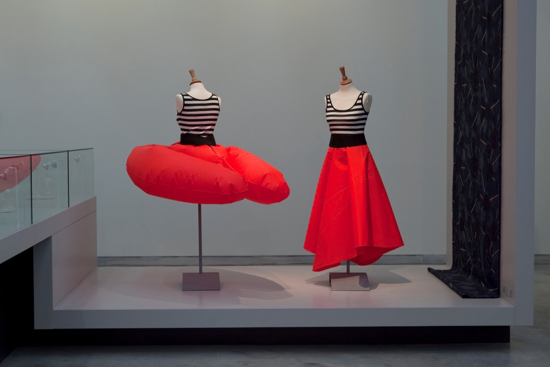 Museum display featuring two dress forms with striped tops and red skirts in a modern gallery setting.