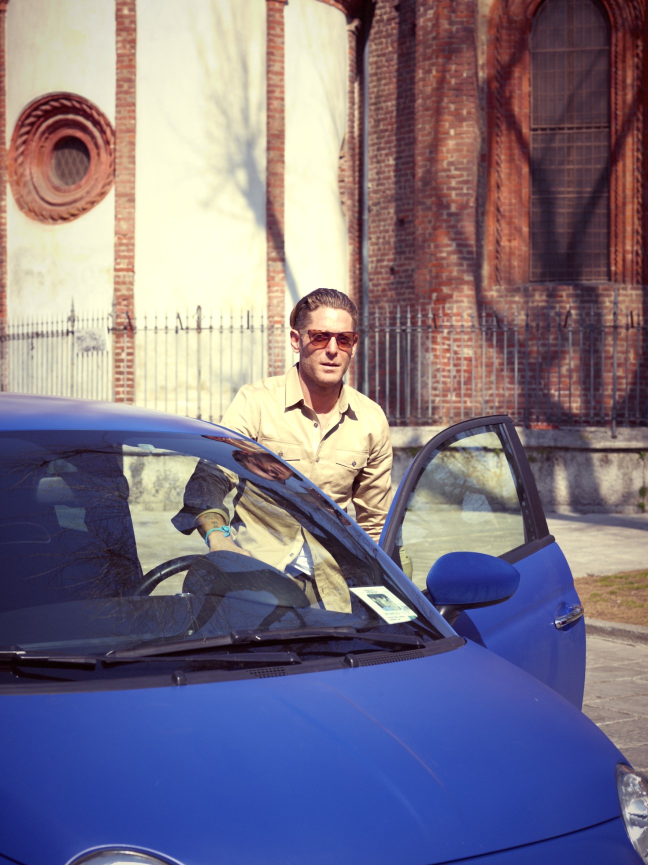 Lapo Elkann stands beside a blue Fiat 500 Abarth in front of a brick basilica in Milan.
