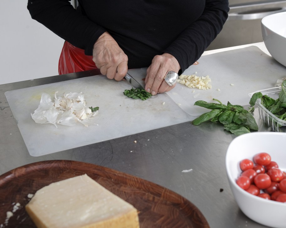 Hands chopping fresh basil on a cutting board with garlic, onion, and tomatoes nearby.