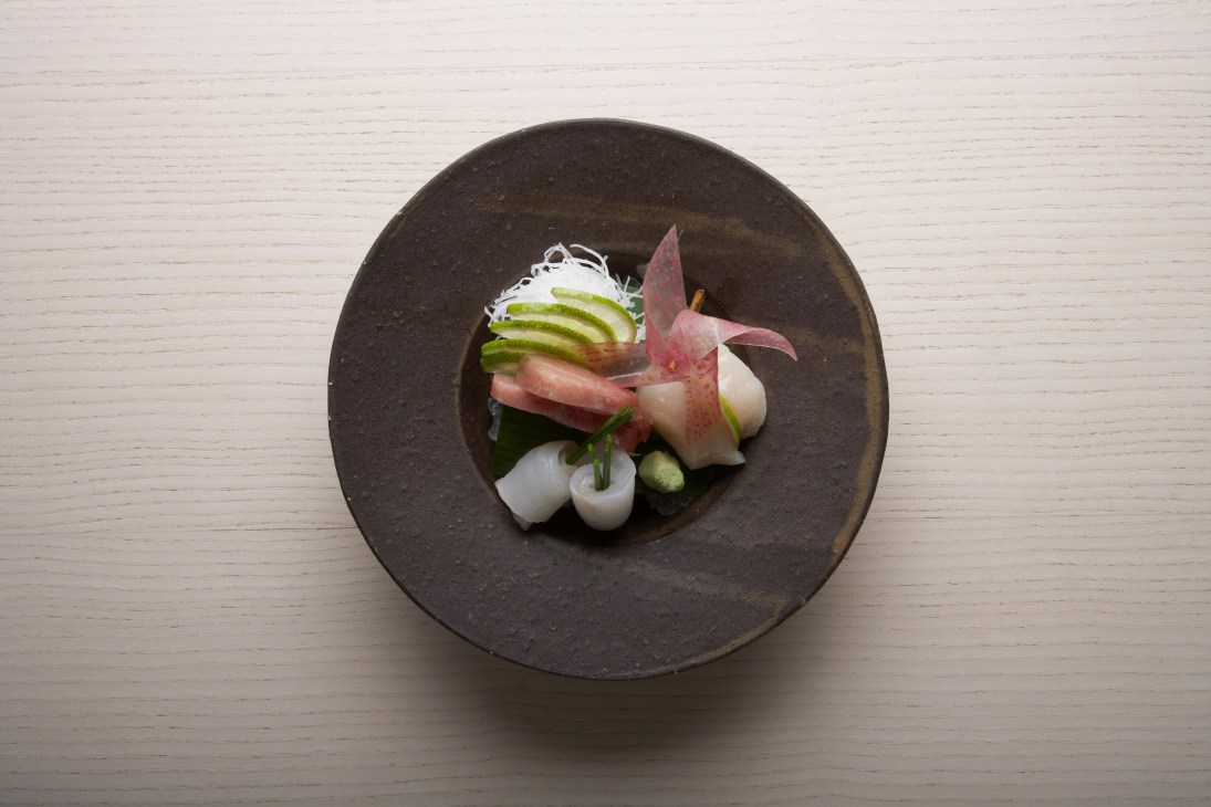 Three kinds of sashimi plated on a dark round plate with shiso leaf garnish.