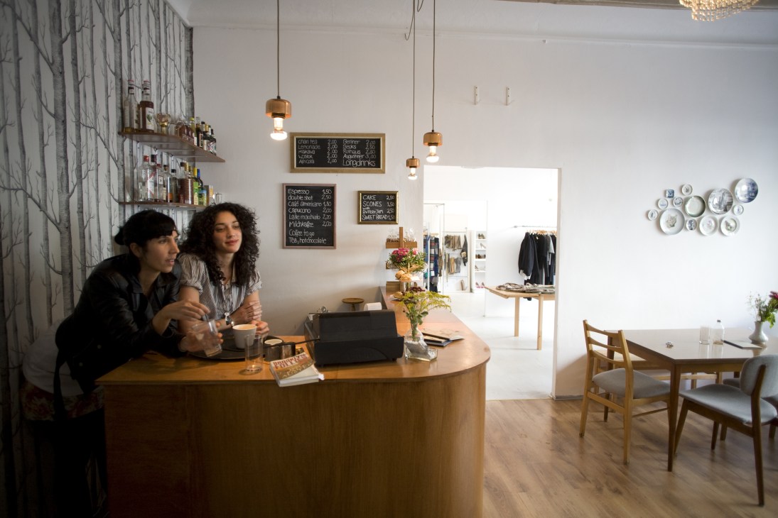 Two women stand at a wooden counter in a bright, minimalist café with menu boards and pendant lights overhead.