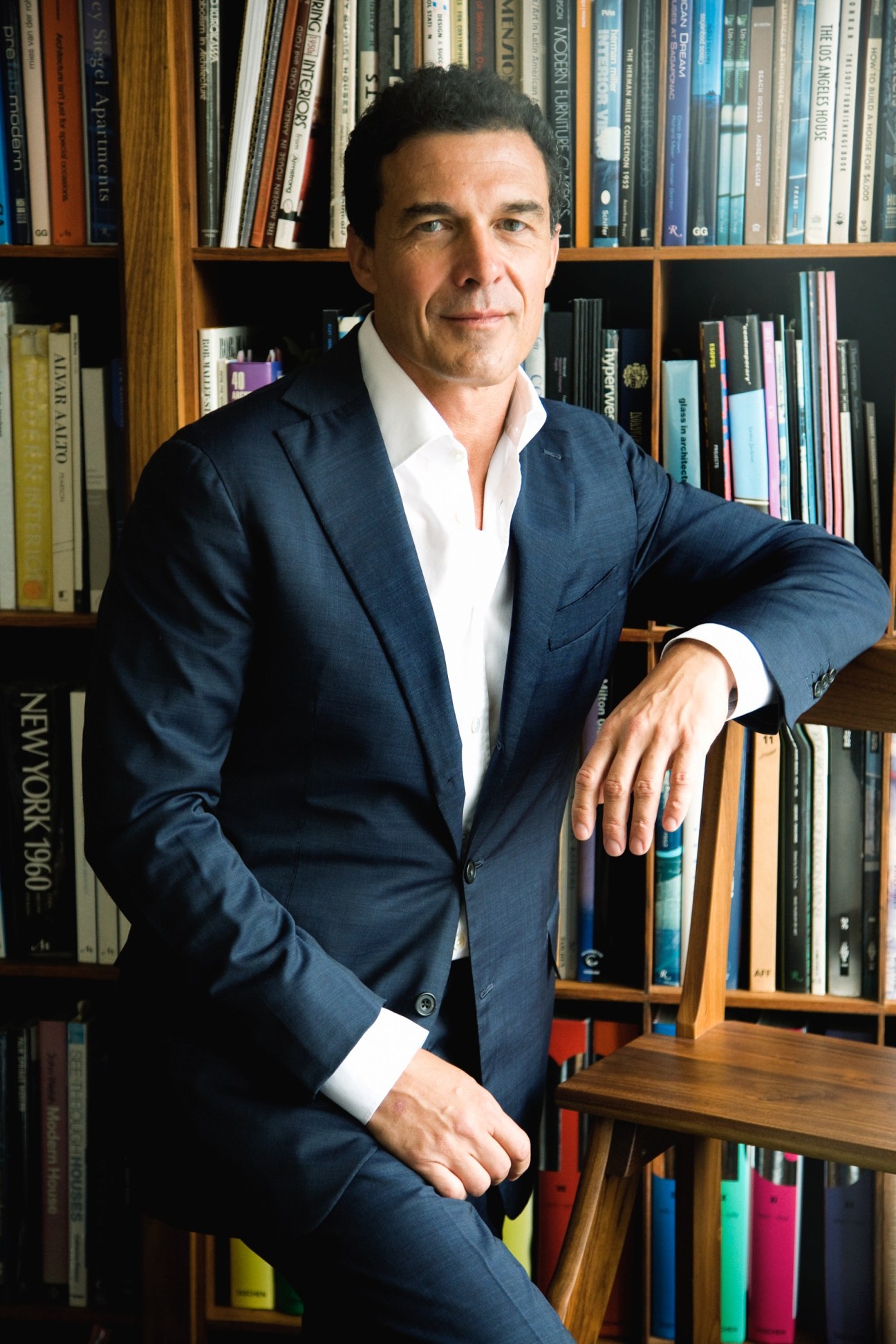Man in navy blazer and white shirt posing against wooden bookshelf