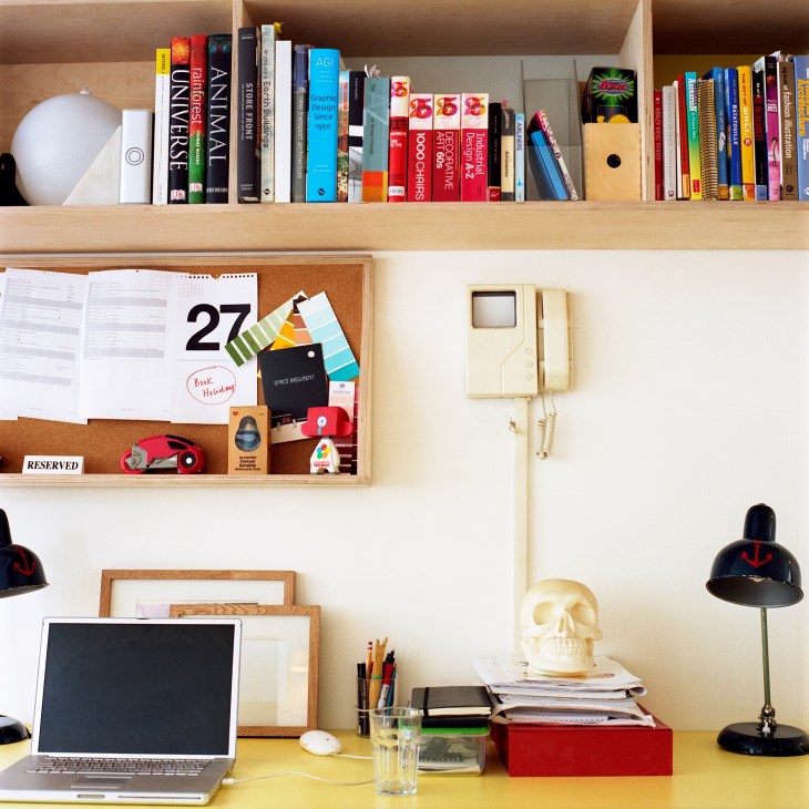 Desk workspace with laptop, yellow desktop, shelving with design books, cork board, vintage telephone, black desk lamp and eclectic decorative objects.