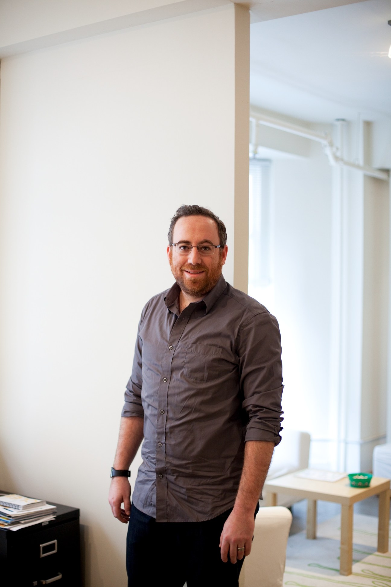 Man wearing glasses and dark button-up shirt standing in bright office space.