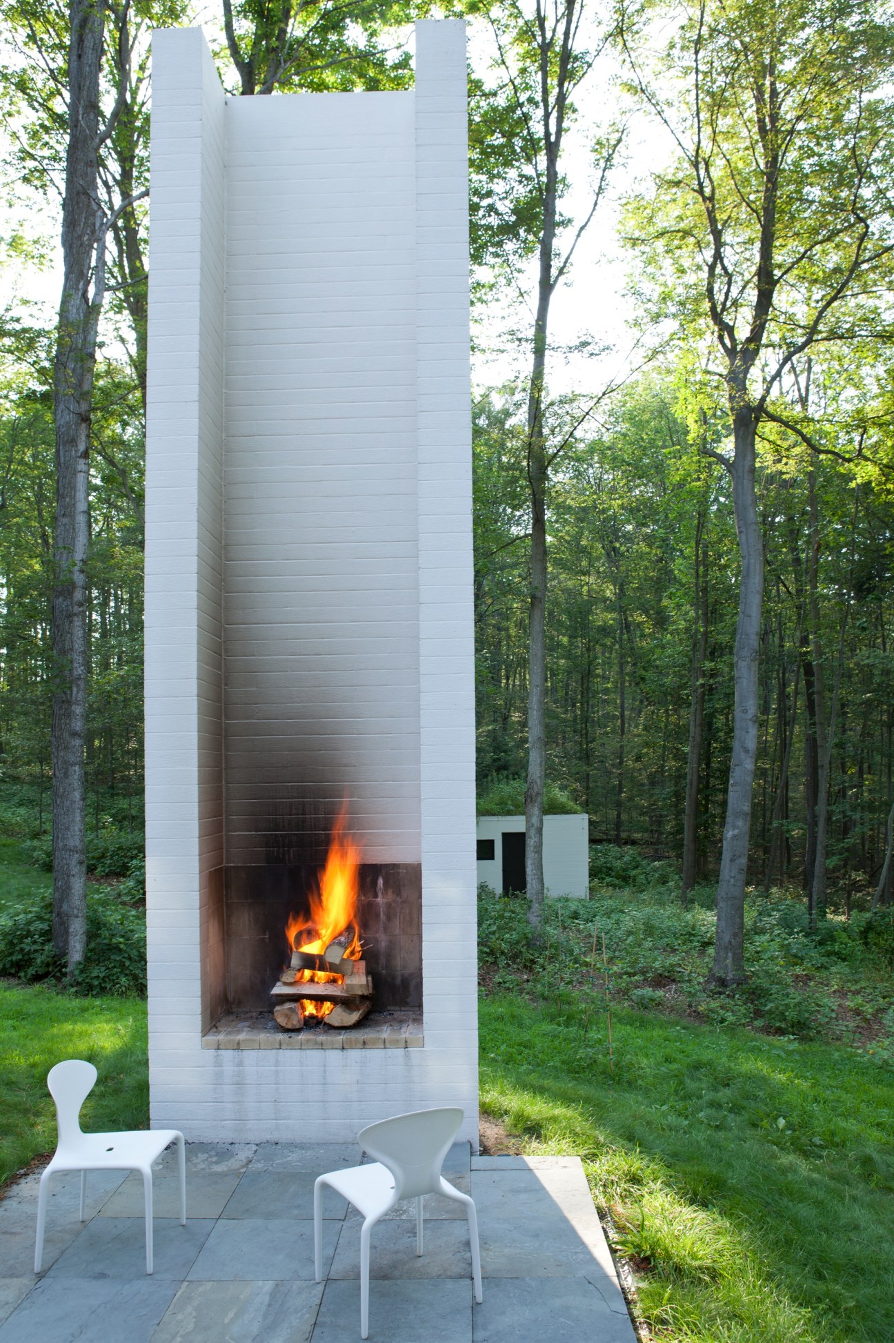 Tall white brick fireplace with active flames surrounded by forest, featuring modern white chairs on stone patio.