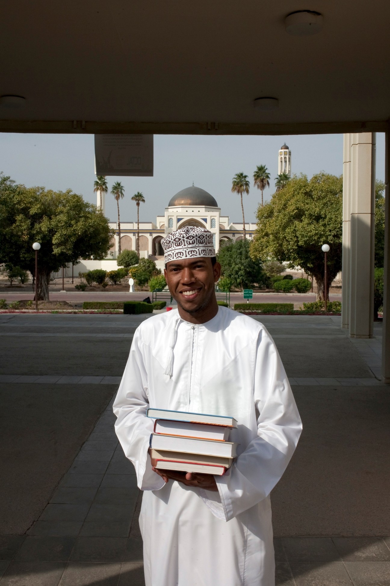Student at Sultan Qaboos University holding books, wearing traditional Omani dress in front of a mosque with dome and minaret.
