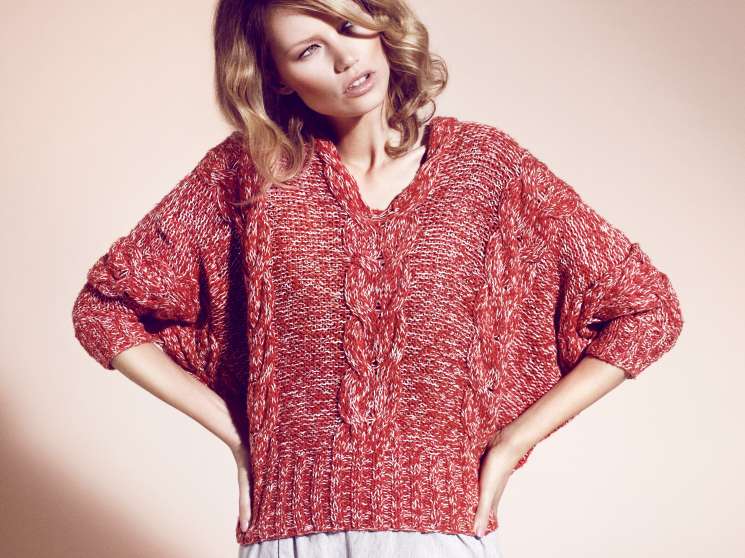 Woman wearing a red cable-knit jumper by Macphee and white shorts by Duša against a neutral background.