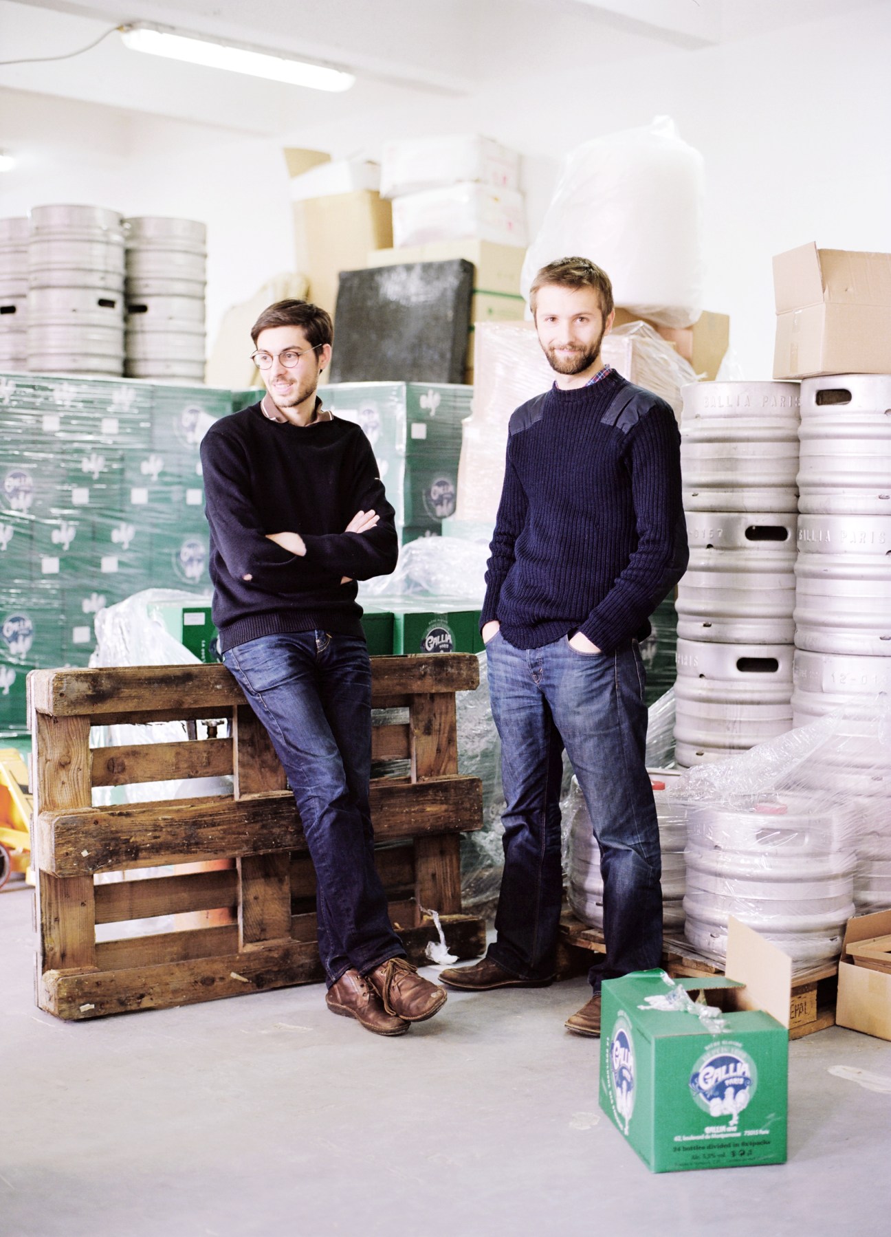 Two men standing in a brewery warehouse surrounded by beer kegs and boxes