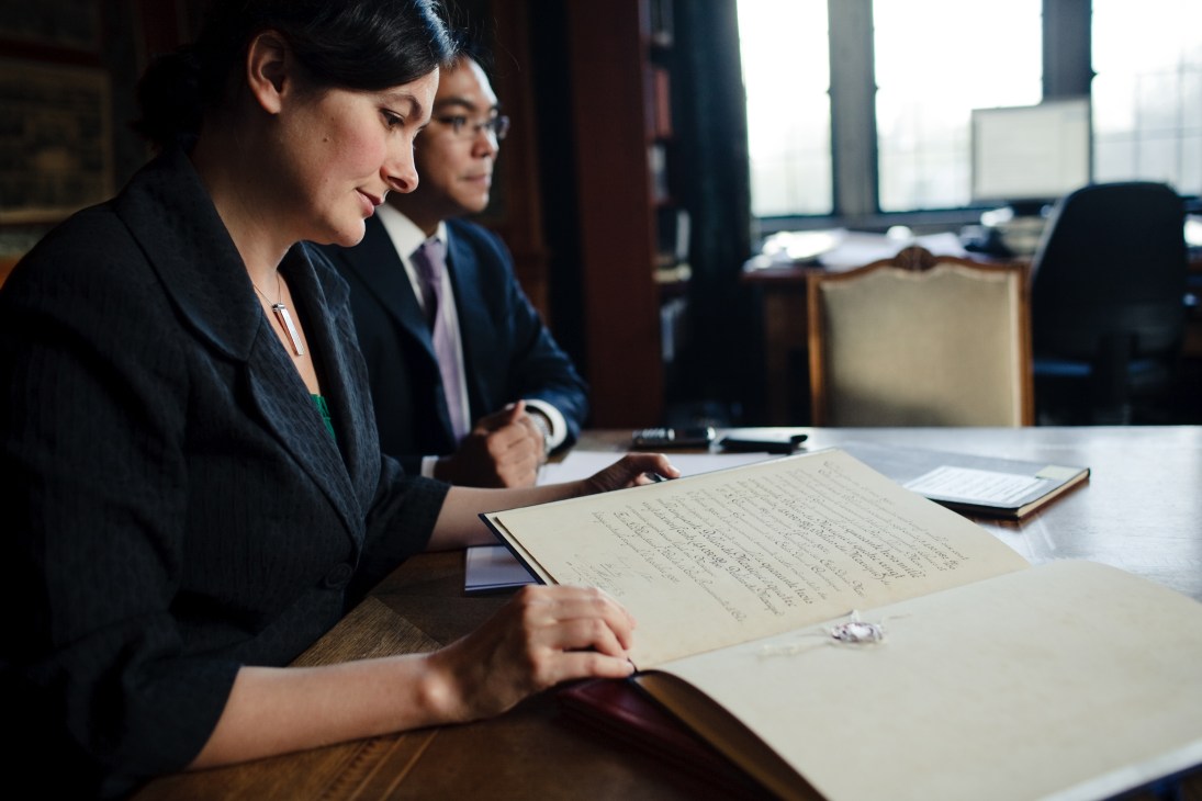 Two lawyers in professional attire review legal documents at a wooden desk in an office.